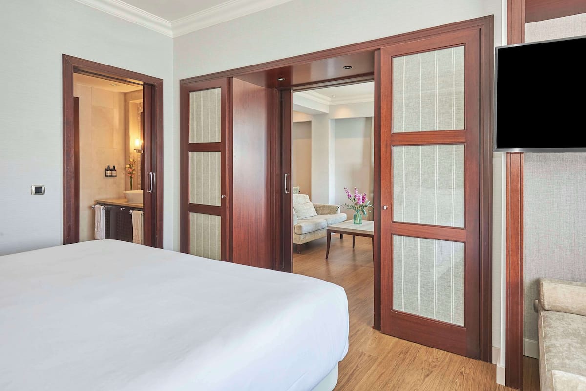 Una habitación de hotel con una cama grande, puertas de madera, un televisor y un baño visible a través de un acceso abierto.