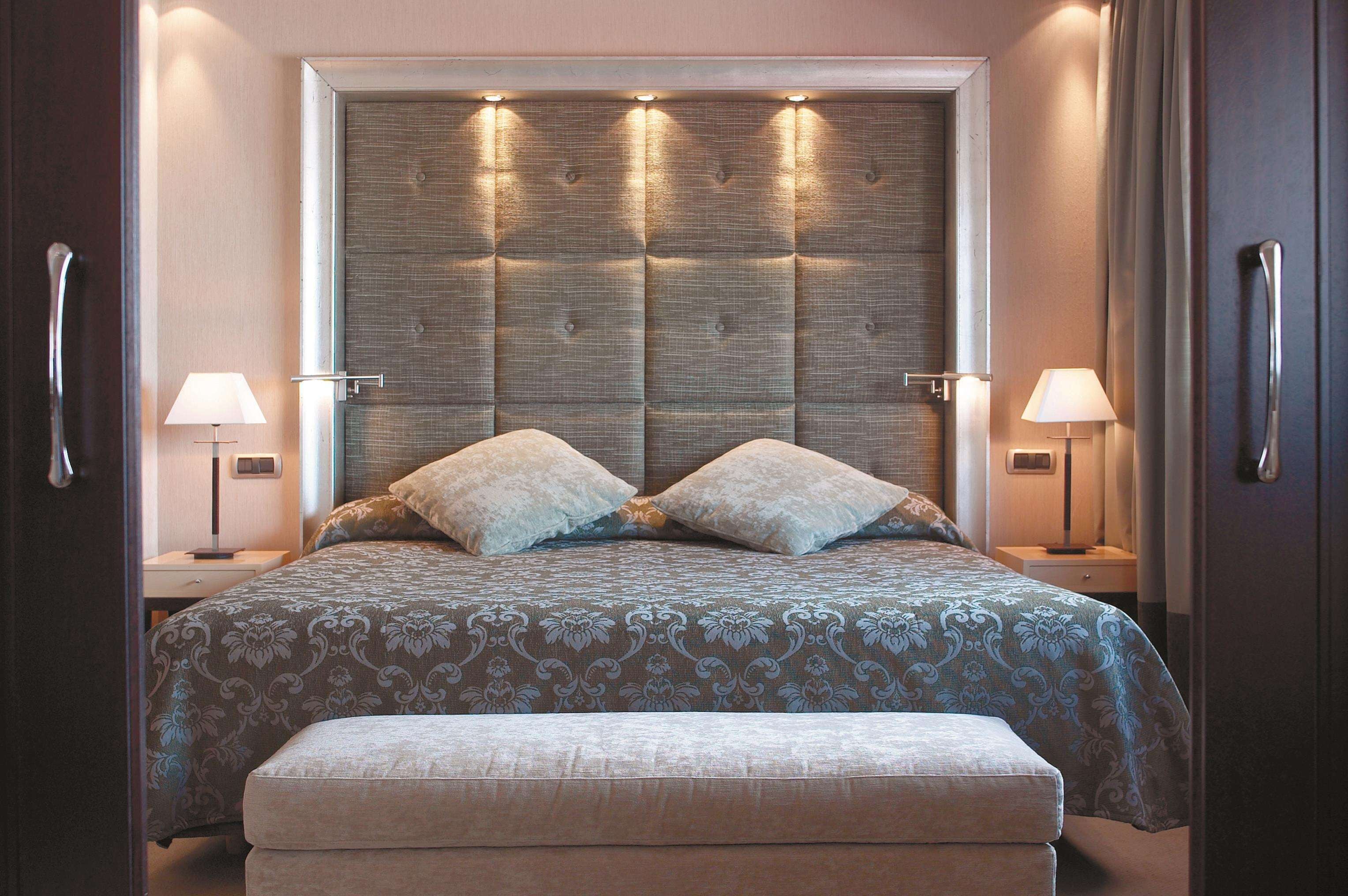 Una cama bien hecha con un cabecero capitonado, dos lámparas de noche y un banco al pie de la cama.