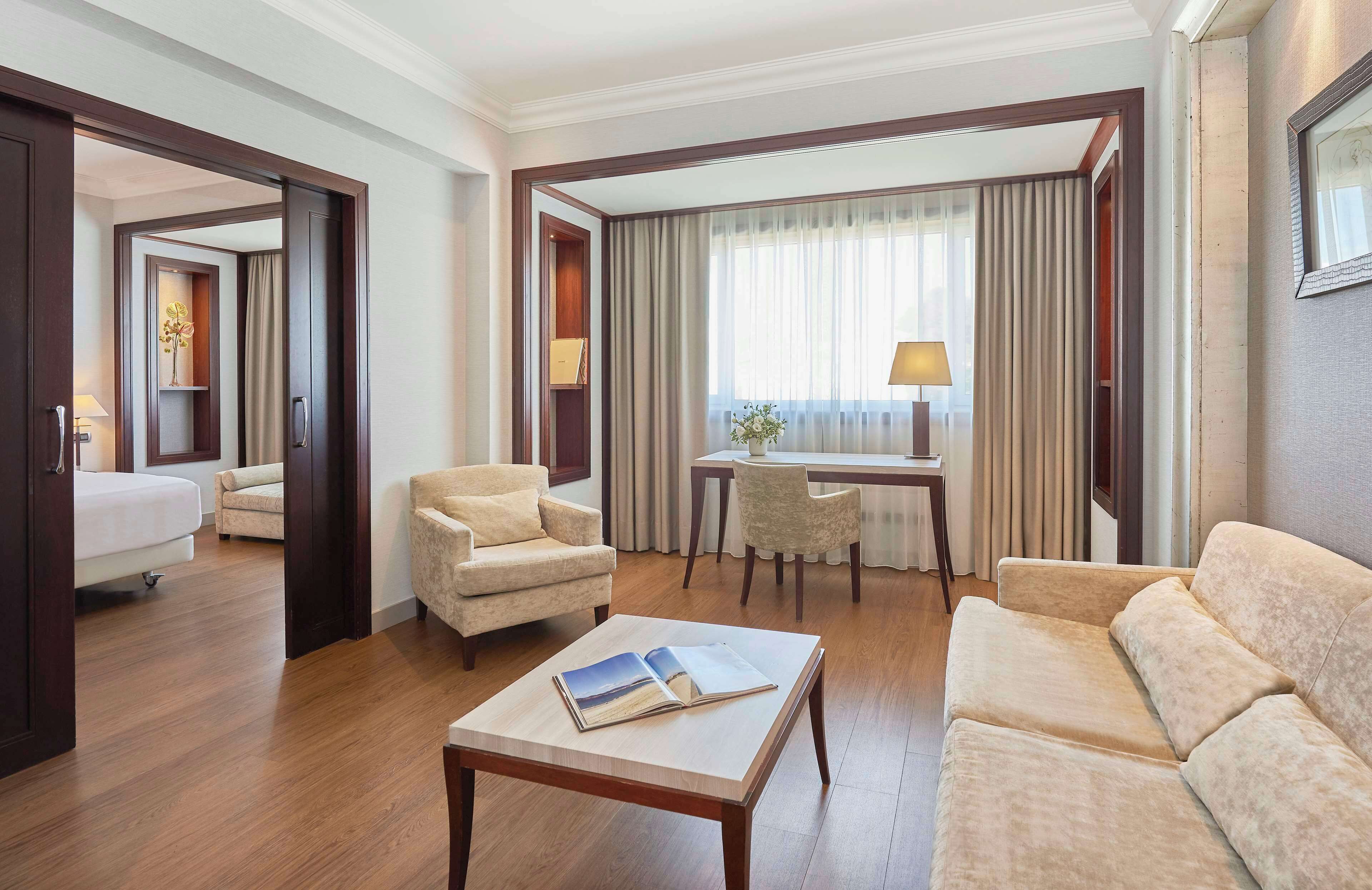 A hotel room with a bed, two armchairs, a sofa, a small table with a book, a desk with a chair, and a window with curtains.