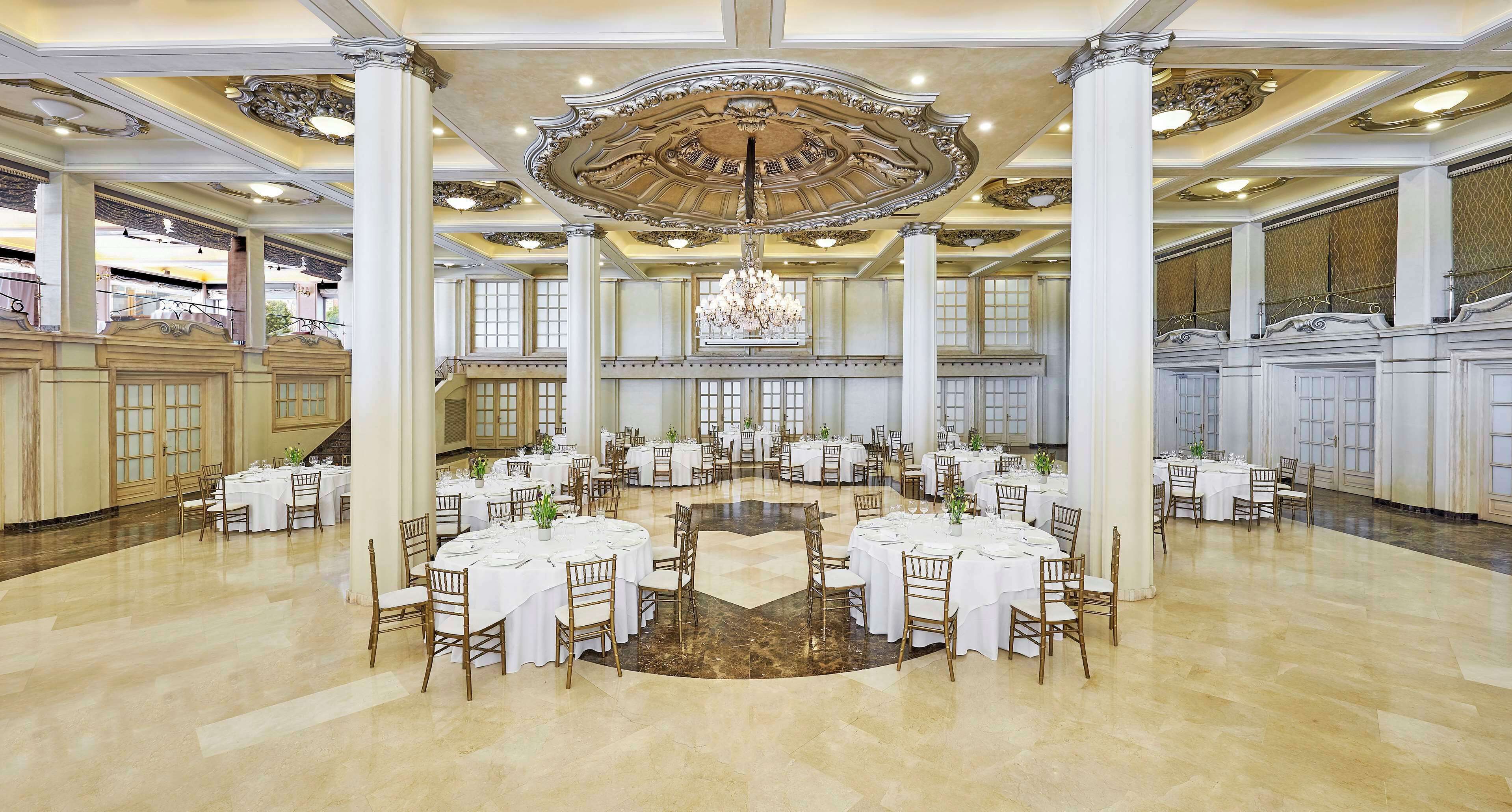 A grand banquet hall with multiple round tables, white tablecloths, and elegant chandeliers.