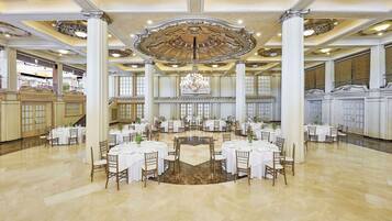 Een statige balzaal met meerdere ronde tafels, witte tafellinnen en elegante kroonluchters.