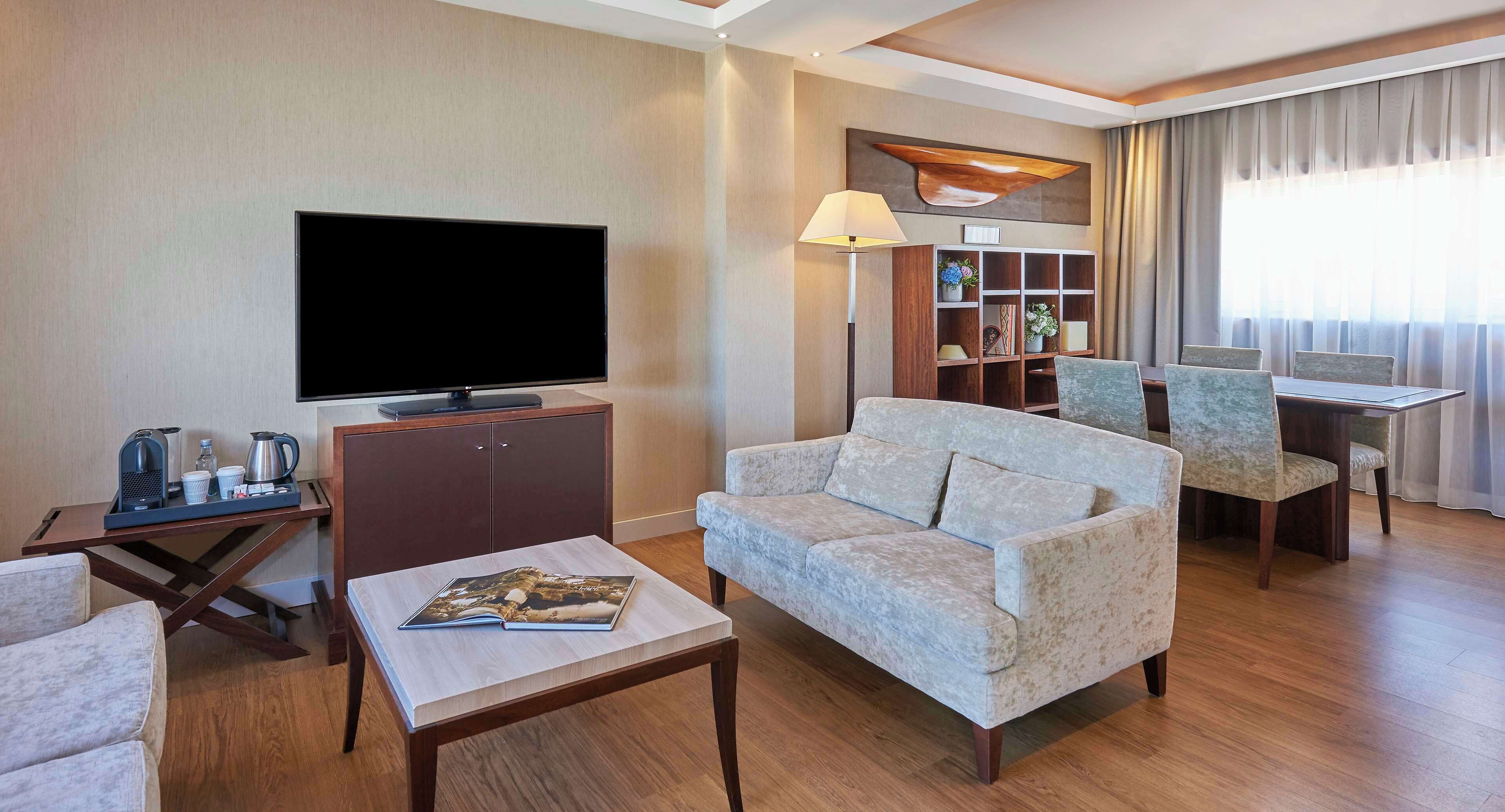 Una habitación de hotel moderna con televisión de pantalla plana, un sofá, una mesa de centro, una estantería y un comedor con sillas.
