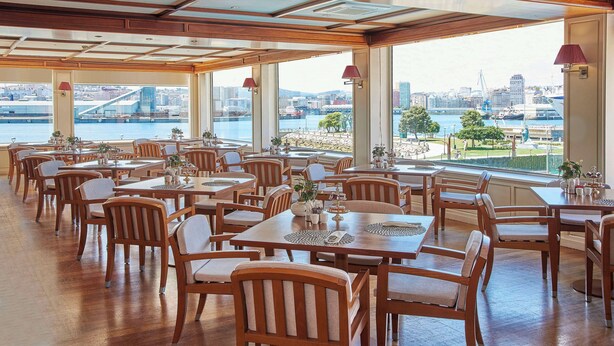 Un comedor amplio con mesas y sillas de madera, ventanas grandes que ofrecen vistas a un paisaje urbano y un río.