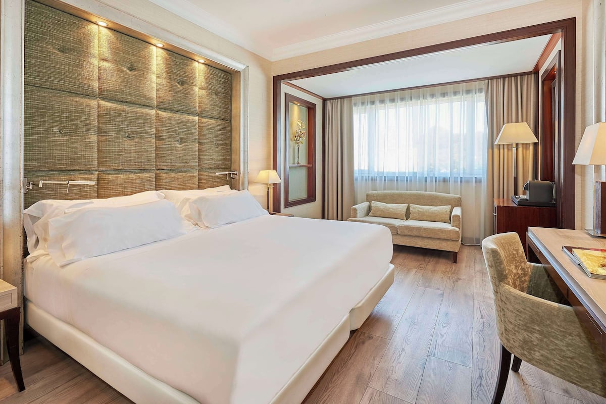 Habitación de hotel con una cama grande, un sofá, un escritorio con lámpara y vistas al exterior a través de la ventana.