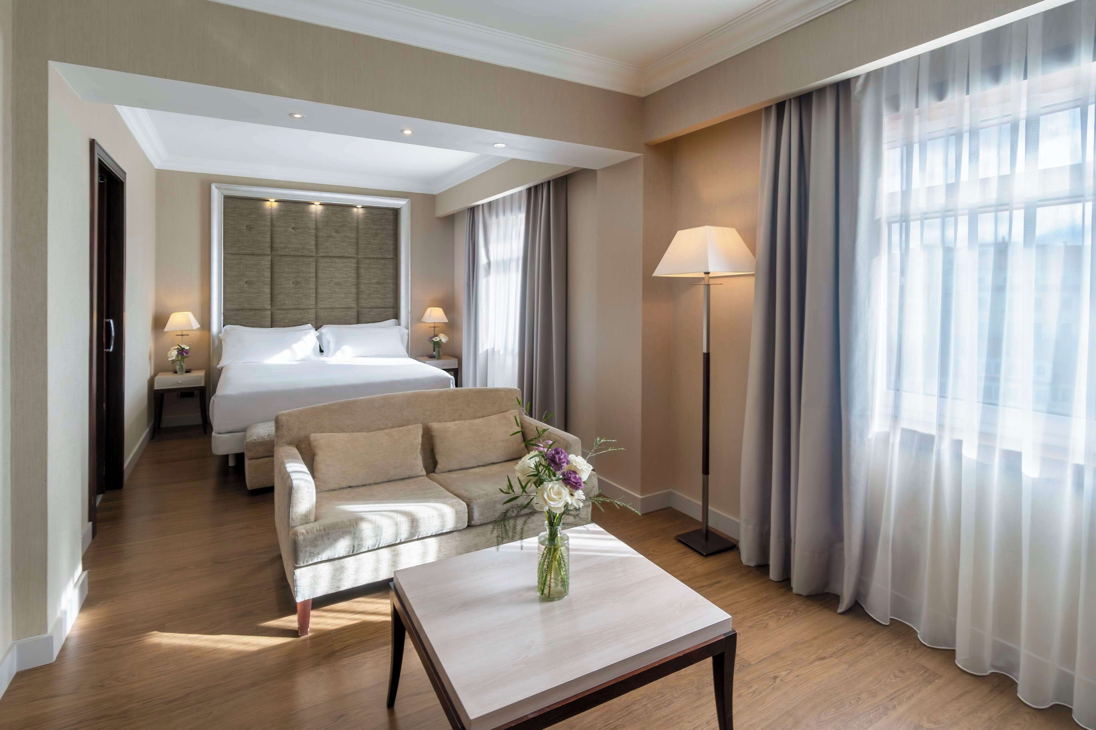 Habitación de hotel con cama, sofá, mesa de centro con un jarrón de flores, lámpara de pie y un ventanal con cortinas.