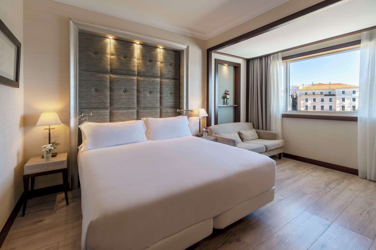 Habitación de hotel con una cama grande, zona de estar con sofá, mesita de noche con lámpara y ventana con vista a la ciudad.