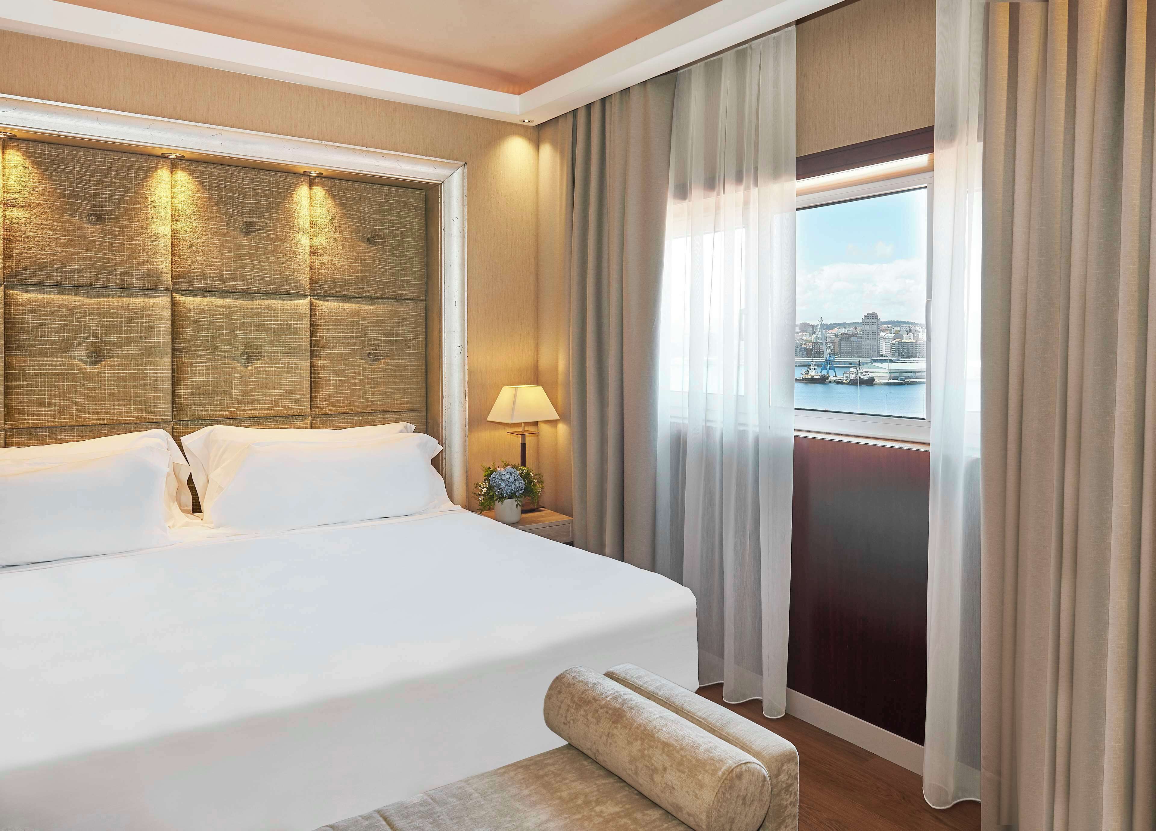 Una habitación de hotel con una cama grande, una mesita de noche con lámpara y vistas a un paisaje urbano a través de la ventana.