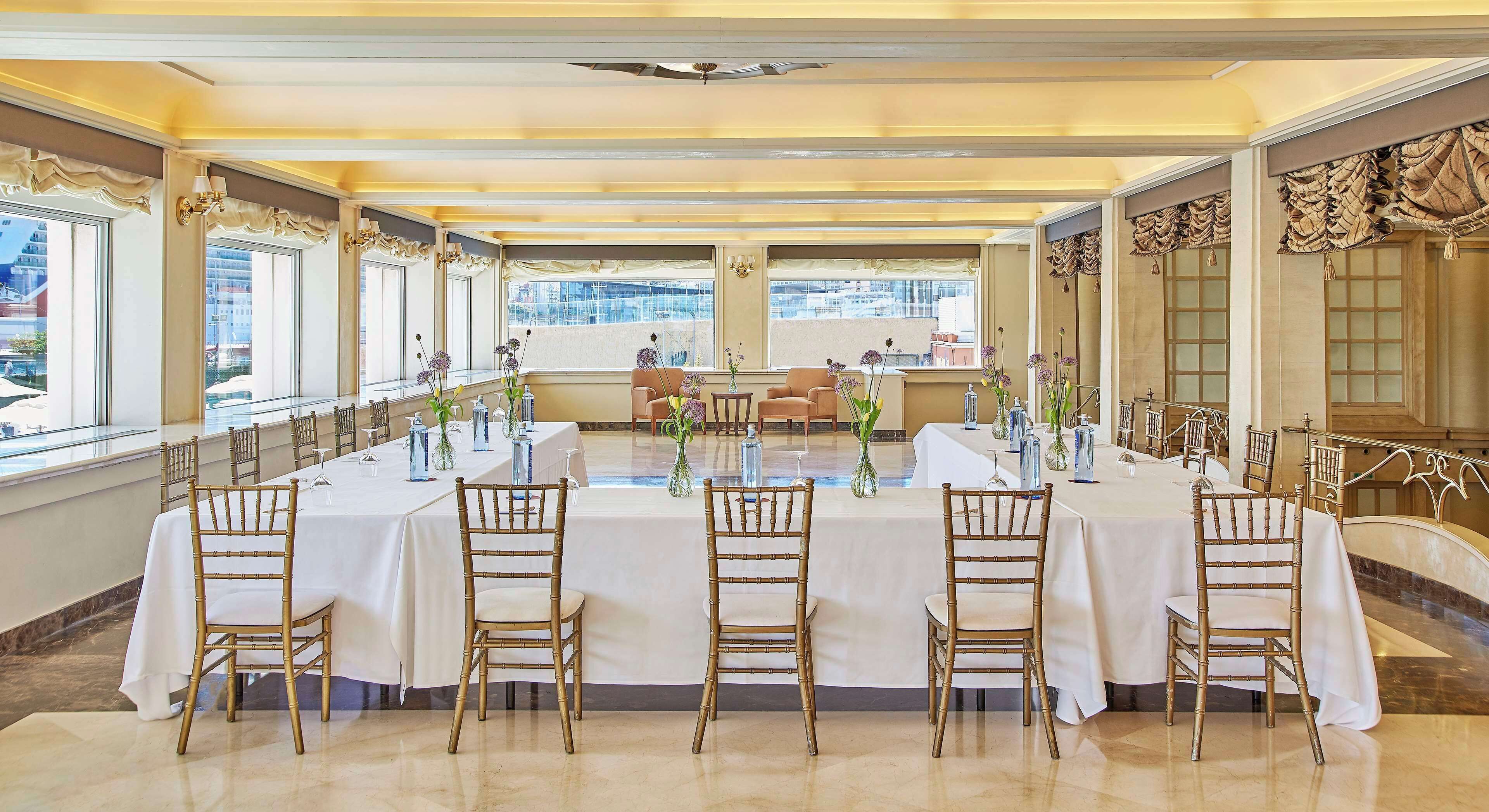 A banquet hall with a long table set for a formal event, flanked by chairs and adorned with floral centerpieces.