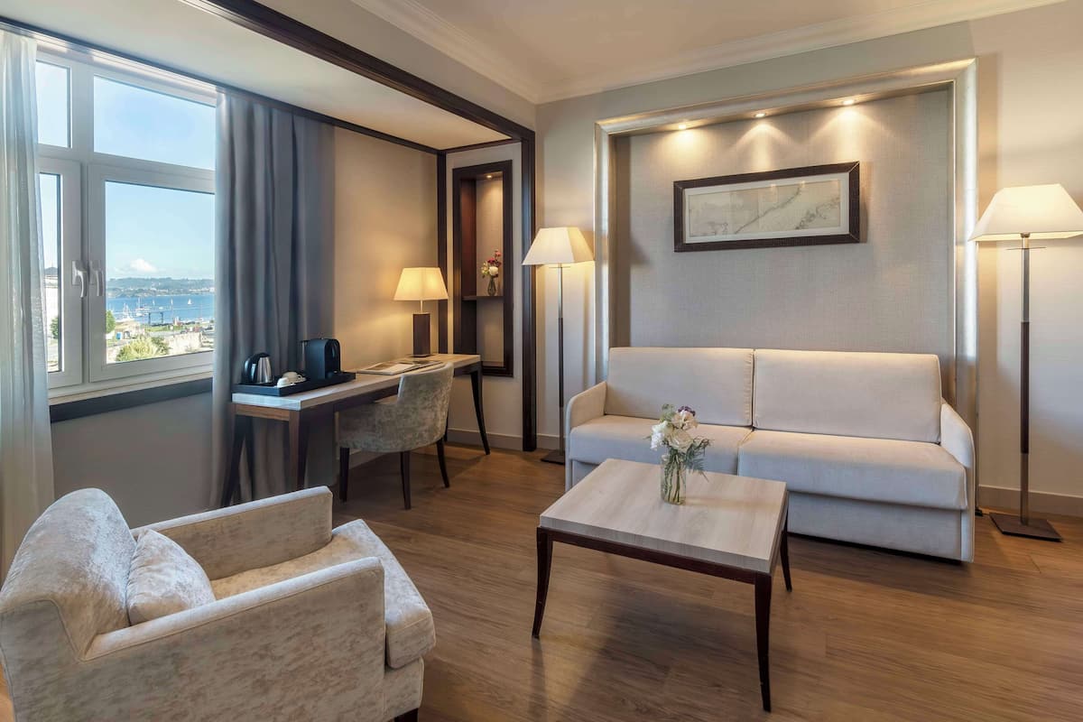 Habitación de hotel con sofá, sillón, escritorio y mesa de centro. Hay una ventana con vista al mar y un cuadro en la pared.