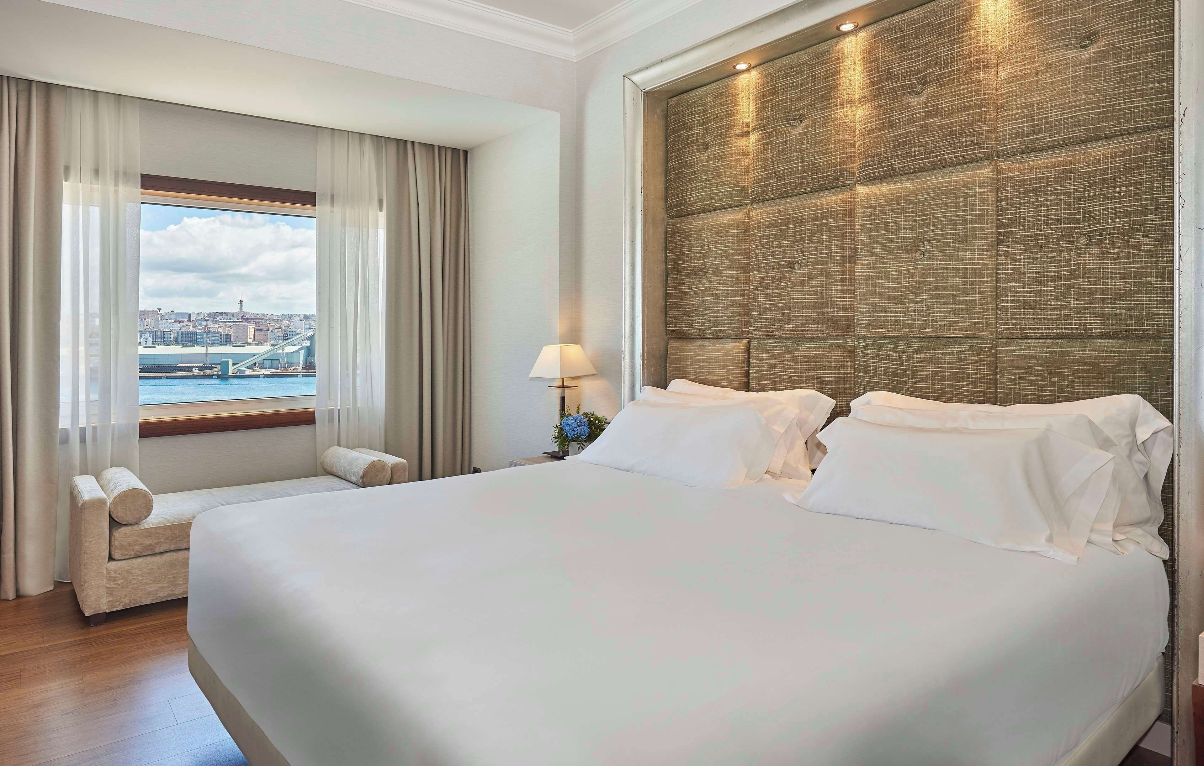 A hotel room with a large bed, a sofa, a window with a view of a cityscape and bridge, and a textured wall panel.