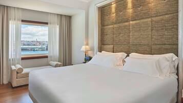 Habitación de hotel con una cama grande, un sofá, una ventana con vistas a un paisaje urbano y un puente, y un panel de pared con textura.
