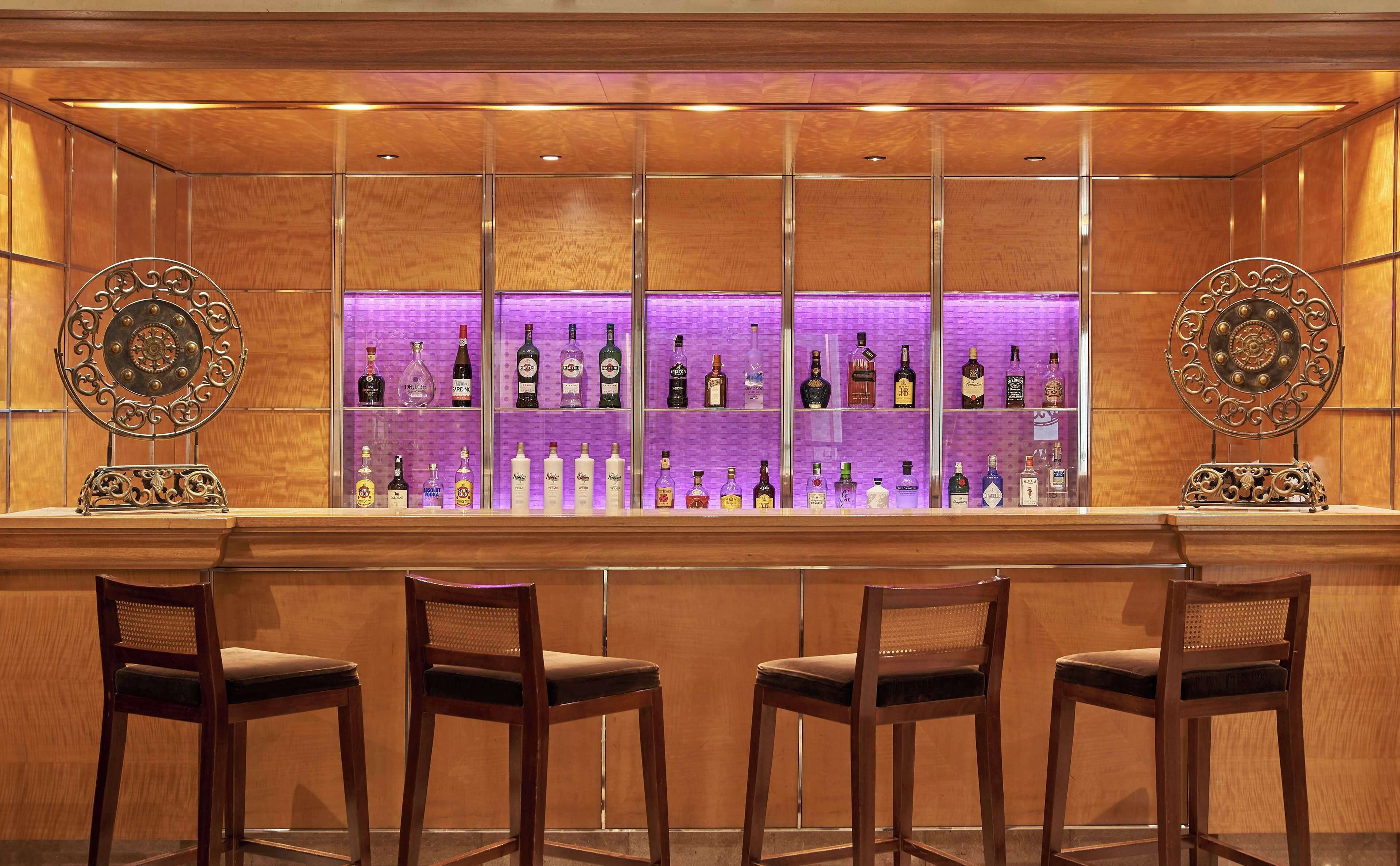 Una zona de bar con paneles de madera y una estantería iluminada en morado que exhibe diferentes botellas de licor.