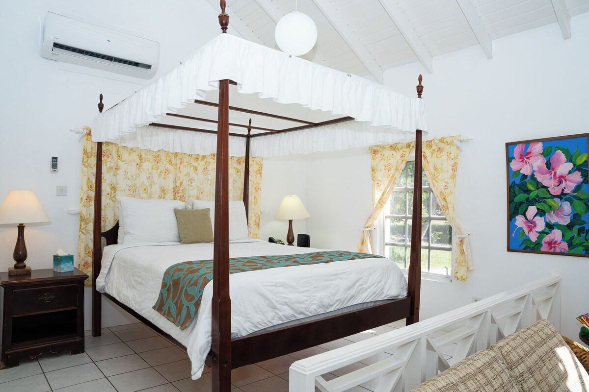 A four-poster bed with floral curtains, a bedside table with a lamp, and a painting of pink flowers on the wall.
