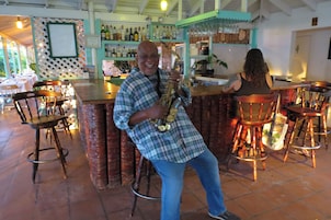 Uma área de bar com cadeiras de madeira, um balcão com garrafas e uma pessoa tocando saxofone.