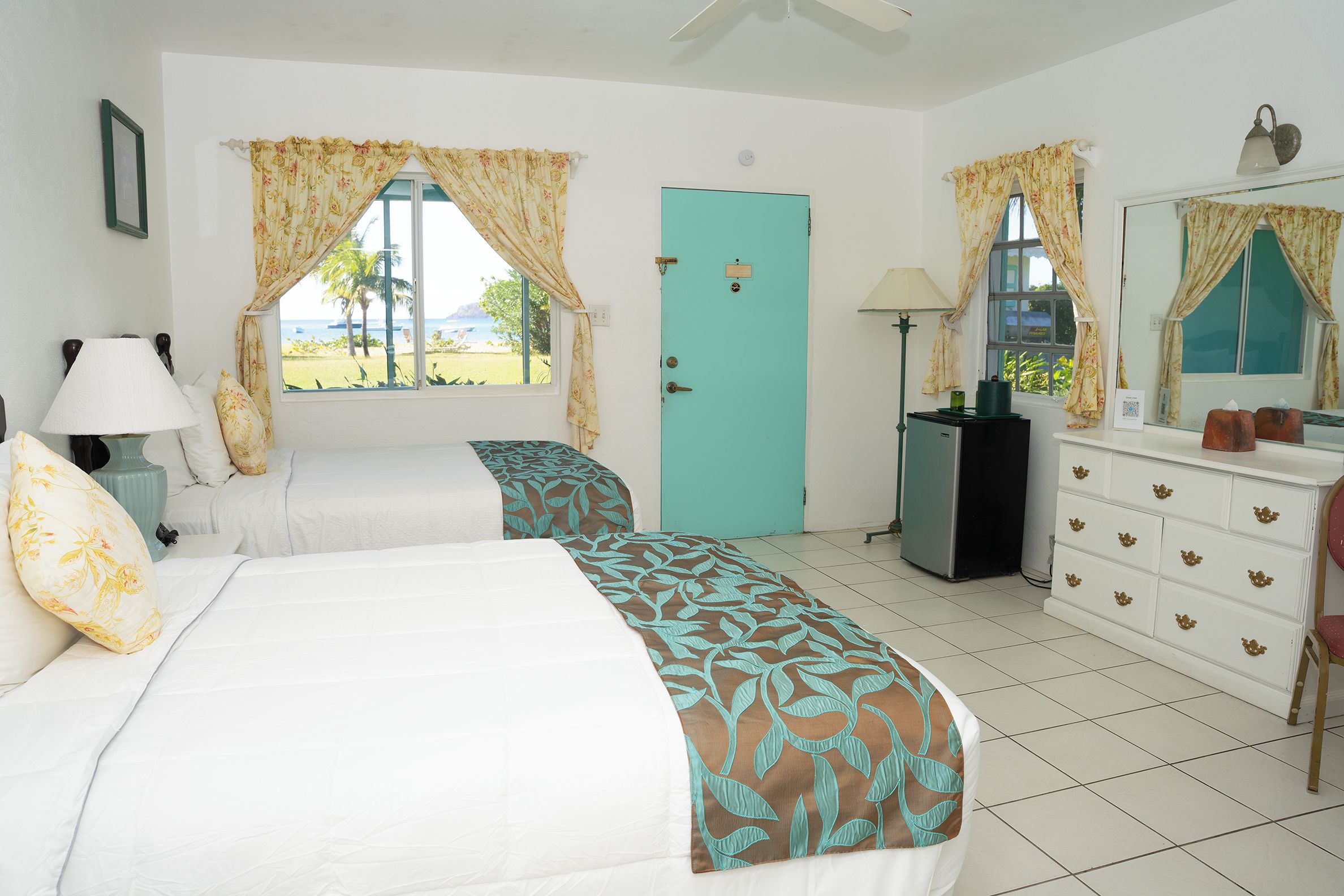 A bedroom with two beds, a door, a dresser, a lamp, and a window with curtains.