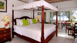 A four-poster bed with white linens, green pillows, and a wooden headboard. There are two bedside tables with lamps, a wooden nightstand, and a view of the beach through large windows. - Oualie Beach Resort (Newcastle)