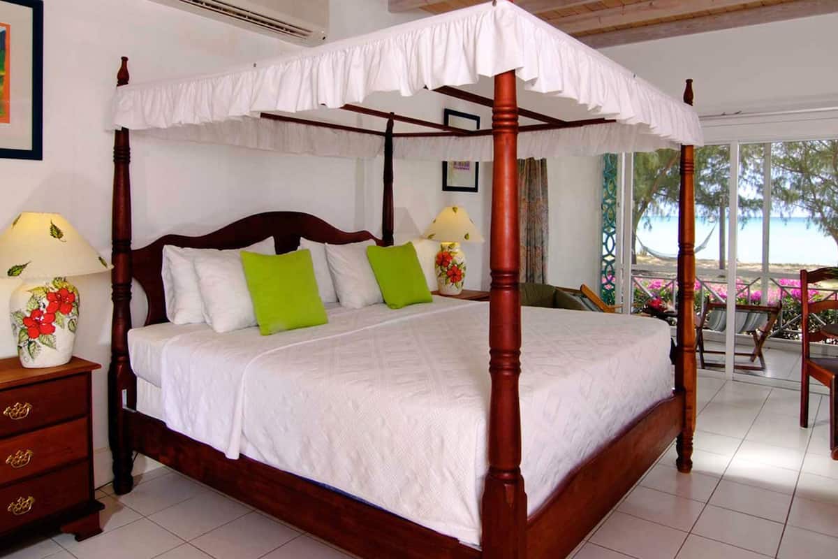 A four-poster bed with white linens, green pillows, and a wooden headboard. There are two bedside tables with lamps, a wooden nightstand, and a view of the beach through large windows.