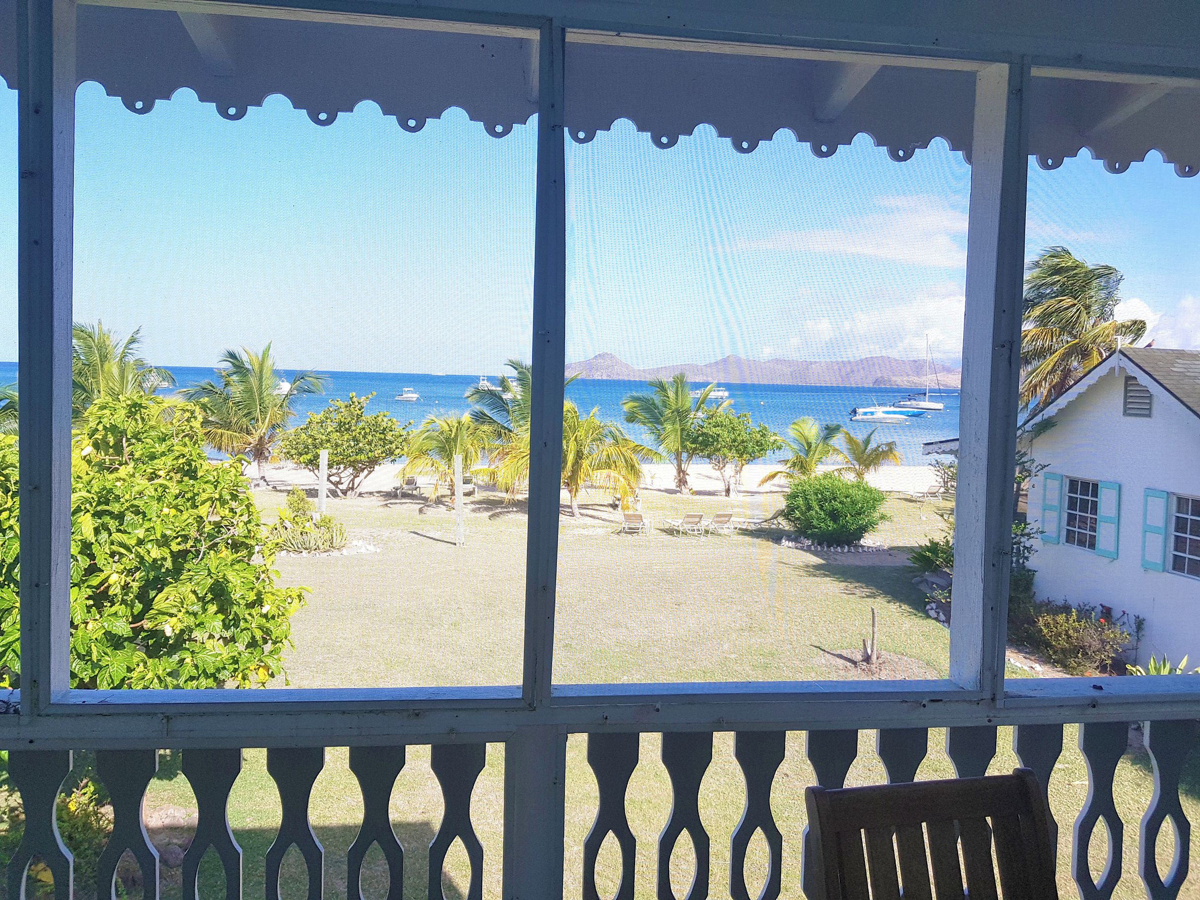 A view from a covered porch with a balcony, overlooking a beach with palm trees and boats.
