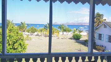 A view from a covered porch with a balcony, overlooking a beach with palm trees and boats.