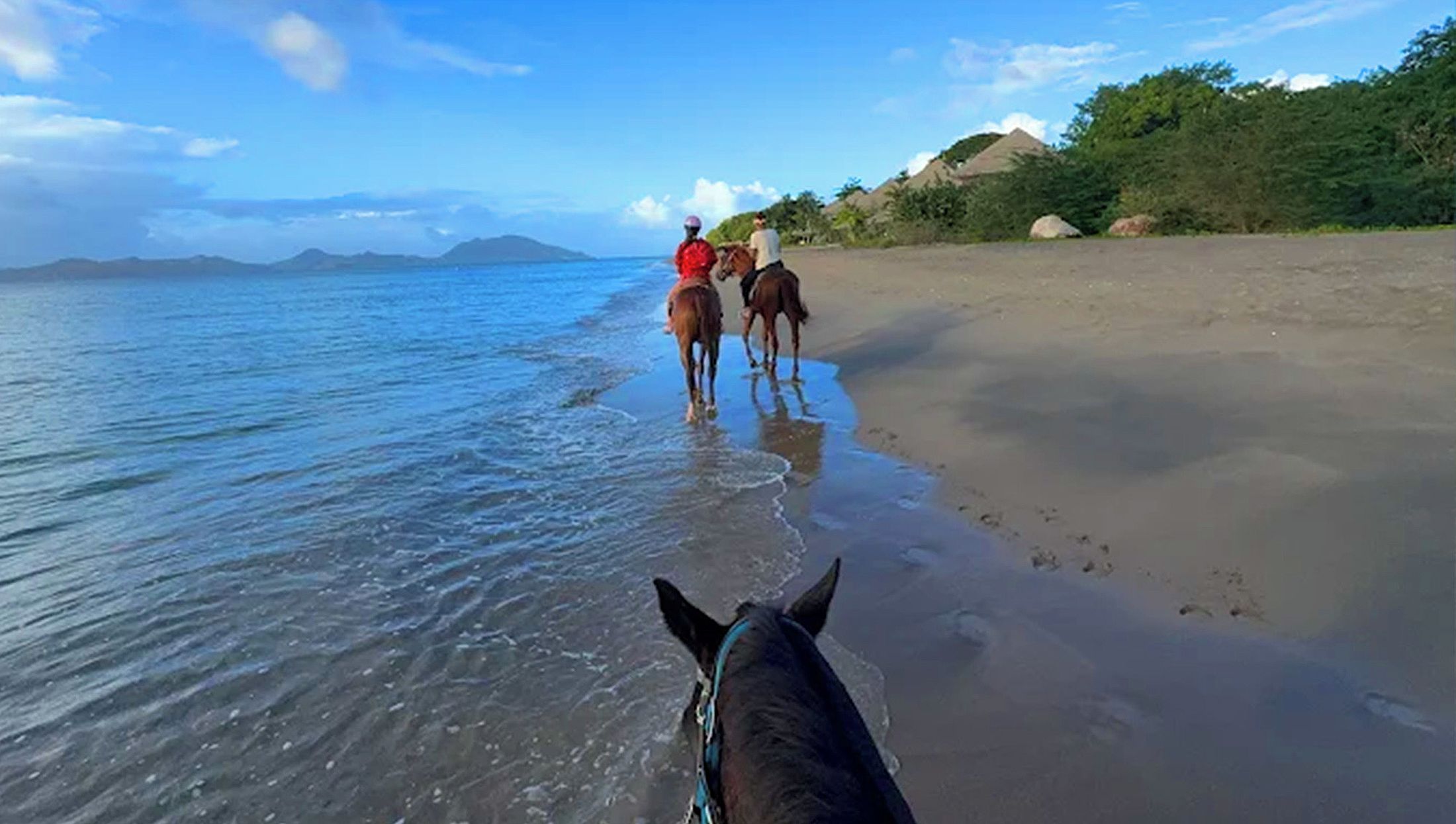 A beach with horses and riders, a clear sky, and distant mountains.