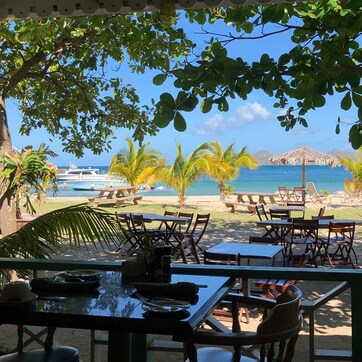 An outdoor seating area with tables and chairs, overlooking a beach with palm trees and a view of the ocean.