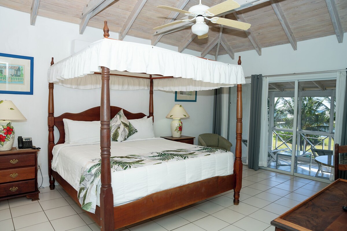 A four-poster bed with a canopy, a wooden nightstand with a lamp, a chair, and a view of the outdoors through a glass door.