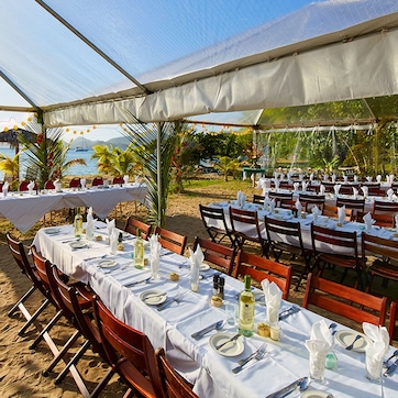 An outdoor dining area with tables set for guests, surrounded by palm trees and a view of the ocean.