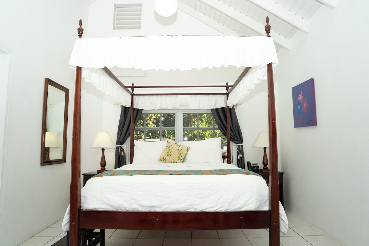 A four-poster canopy bed with a white canopy, two bedside tables with lamps, a nightstand, and a window with curtains.