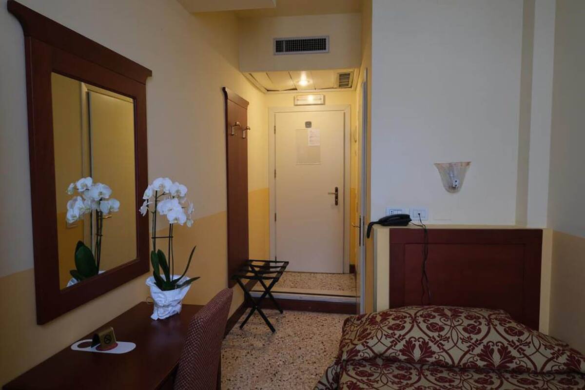 A hotel room with a bed, a bedside table with a vase of white flowers, a mirror, a small table with a tray, and a door leading to a hallway.