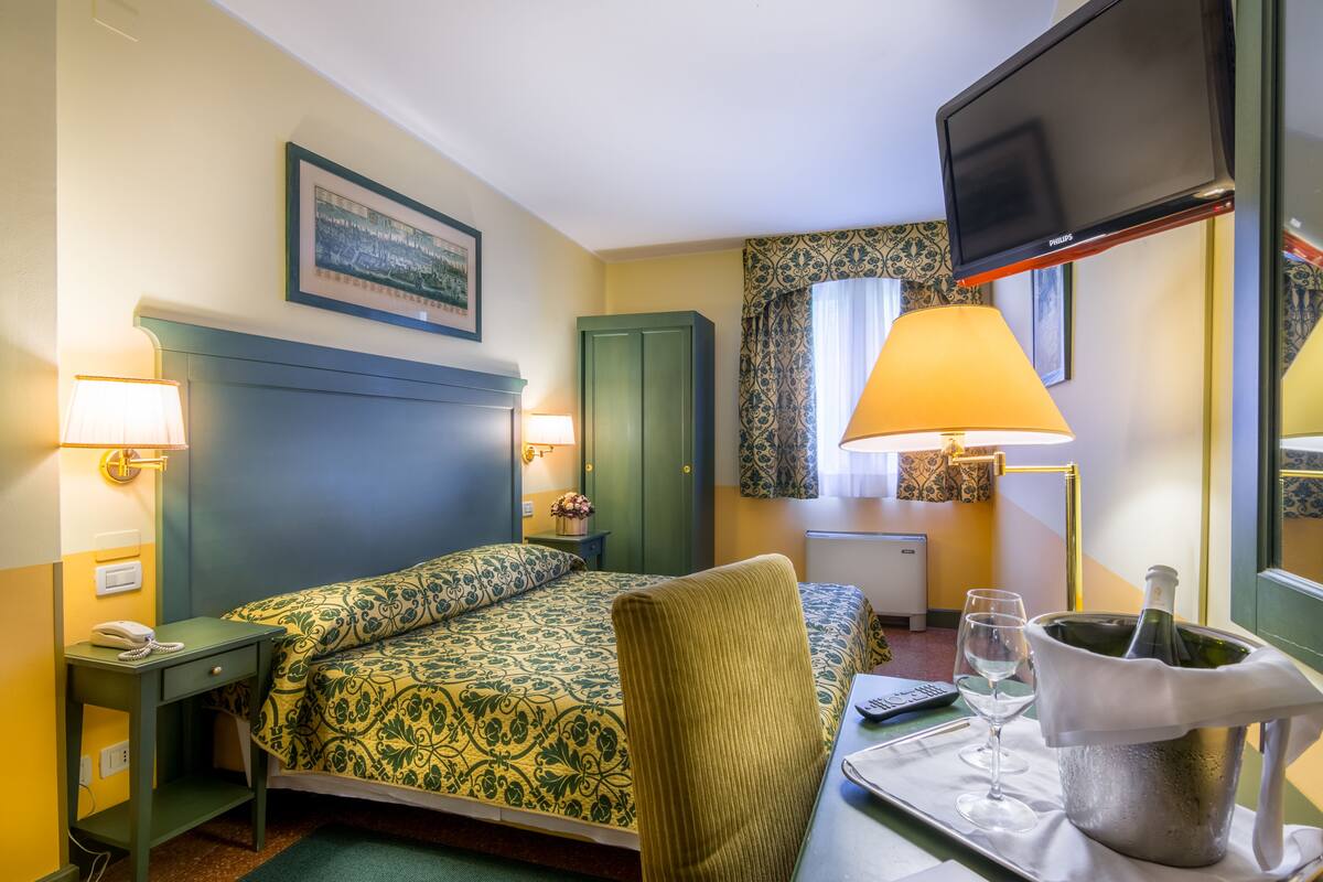 A hotel room with a bed, bedside lamps, a television, a minibar, and a window with curtains.