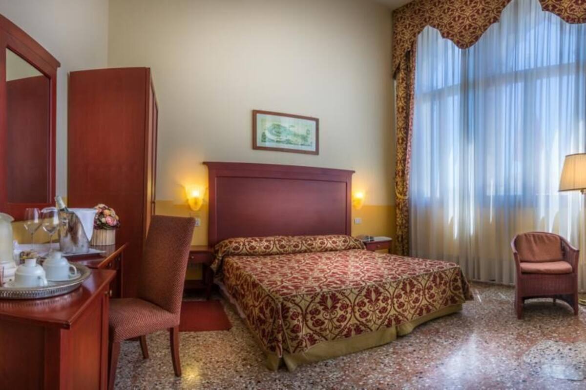 A hotel room with a bed, two chairs, a desk with a lamp, a mirror, and a window with curtains.