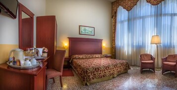 Camera d'albergo con un letto, due poltrone, una scrivania con una lampada, uno specchio e una finestra con tende.