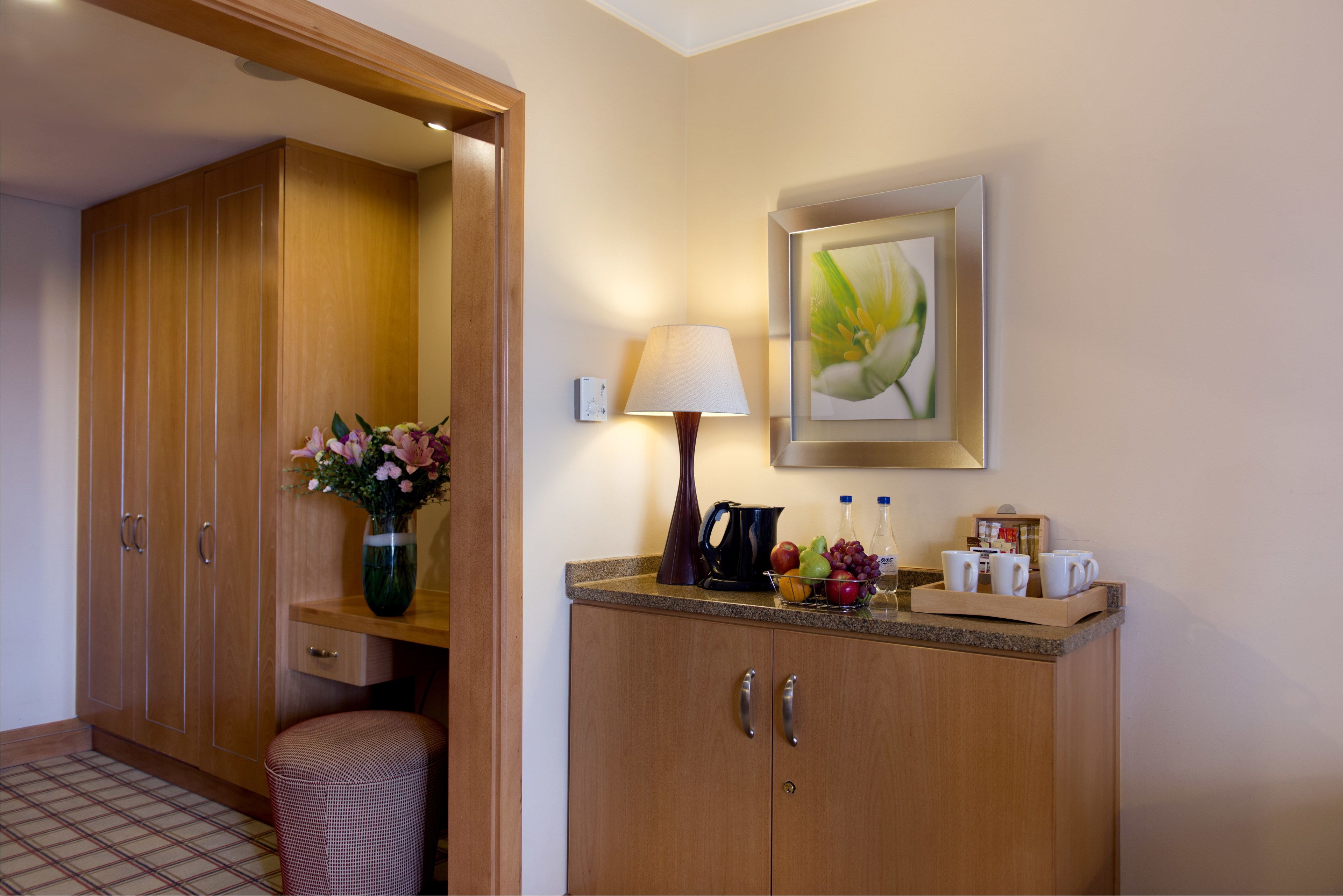 Habitación de hotel con un armario de madera, una mesita con un jarrón de flores, una lámpara y un cuadro en la pared.
