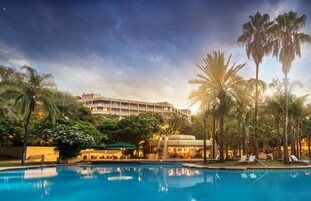 A hotel with a swimming pool, palm trees, and a large building in the background.