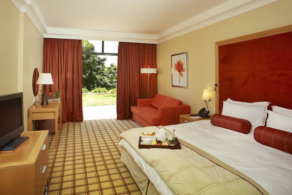 Une chambre d’hôtel comprenant un lit, une télévision, un bureau, une chaise, un canapé, une table de chevet, une lampe et une fenêtre avec des rideaux.