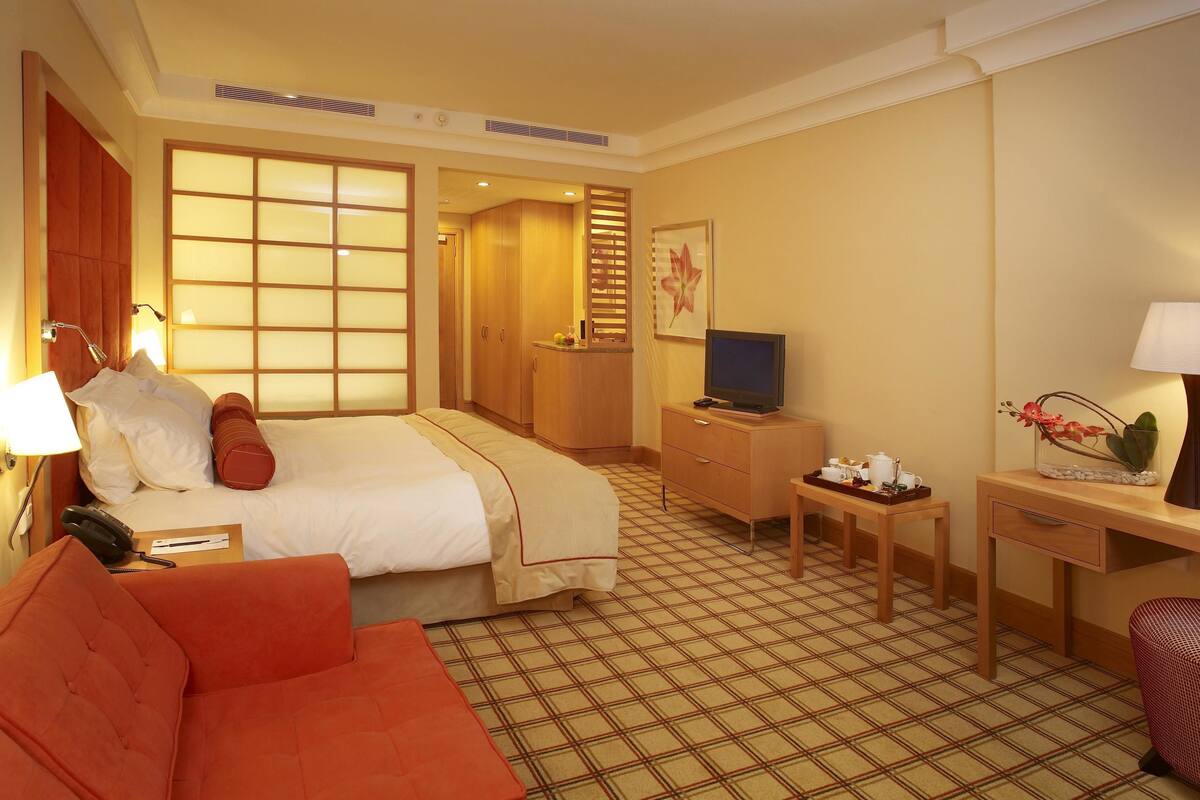Une chambre d’hôtel avec un lit, un bureau, une télévision, une lampe et une petite table sur laquelle est posé un vase de fleurs.