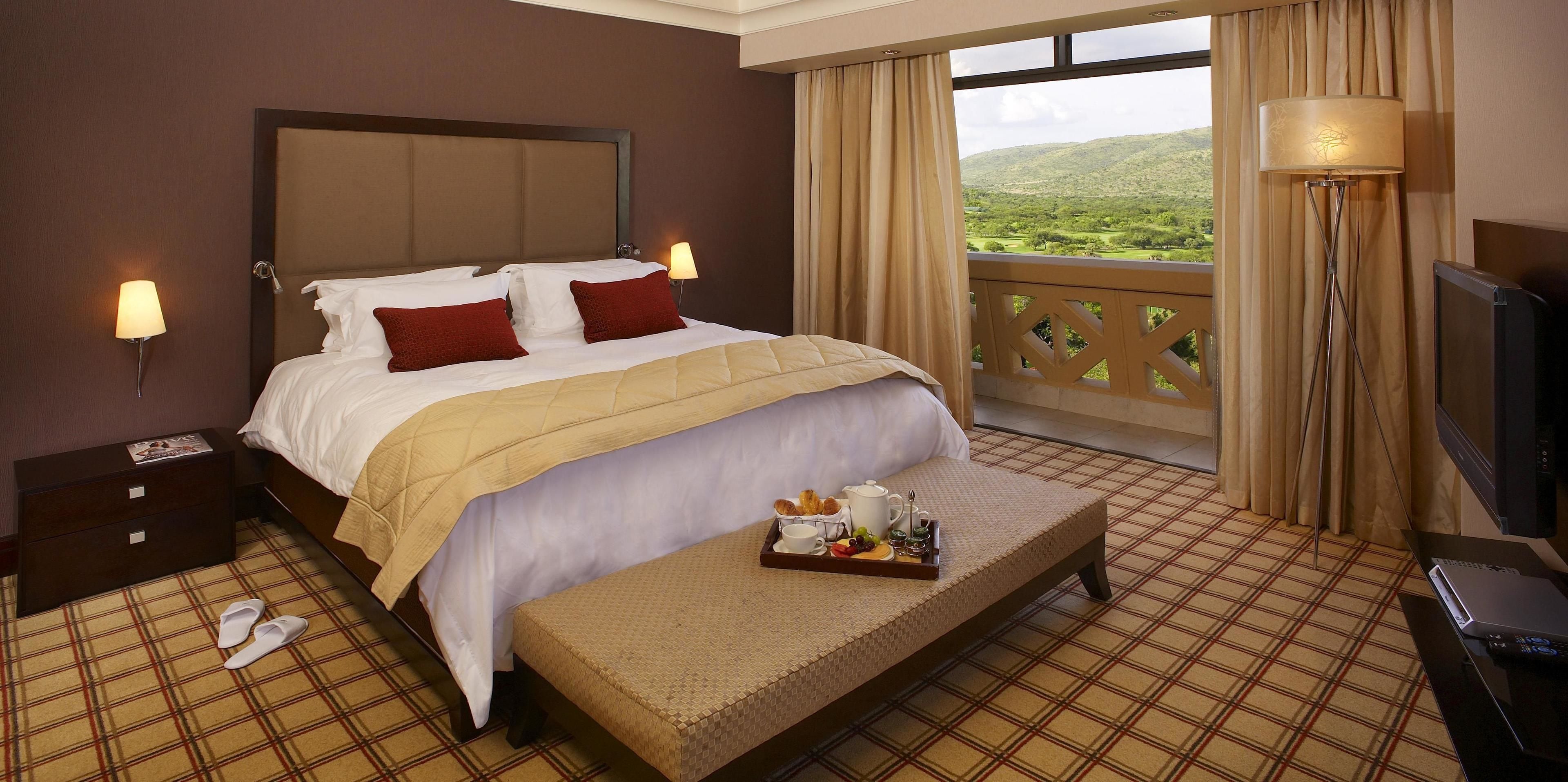Quarto de hotel com cama, luminárias de cabeceira, bandeja com itens de café da manhã, televisão e varanda com vista para área verde.