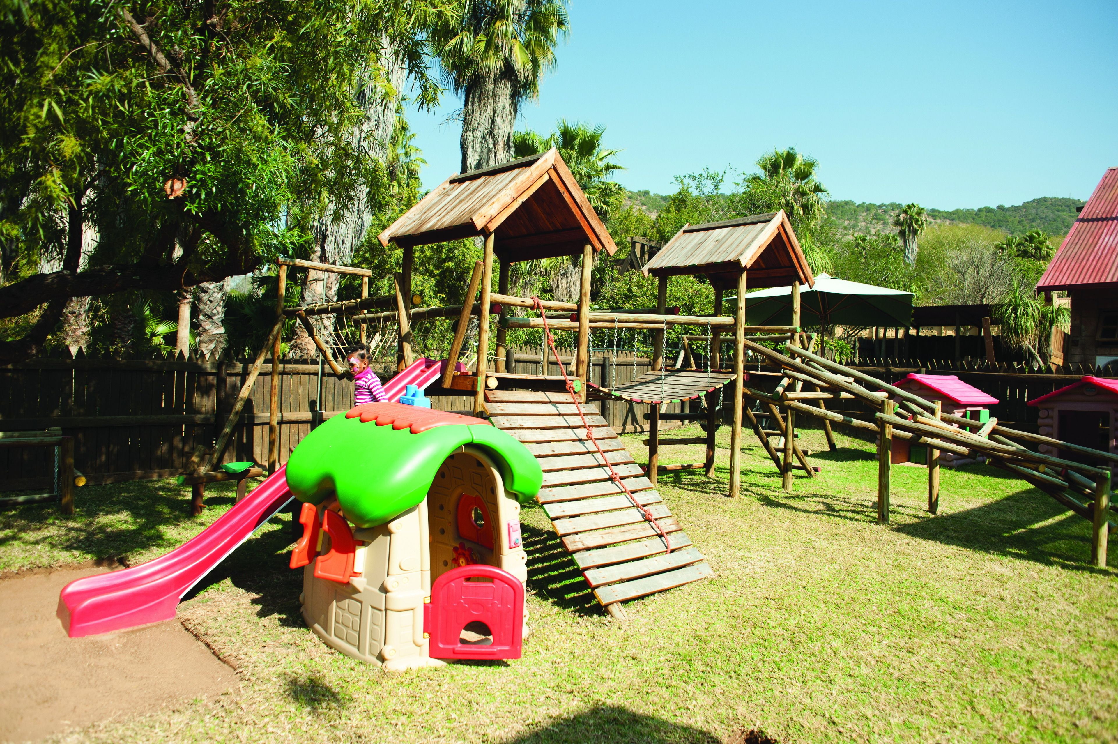 A playground with a slide, climbing structures, and a small house with a green roof.