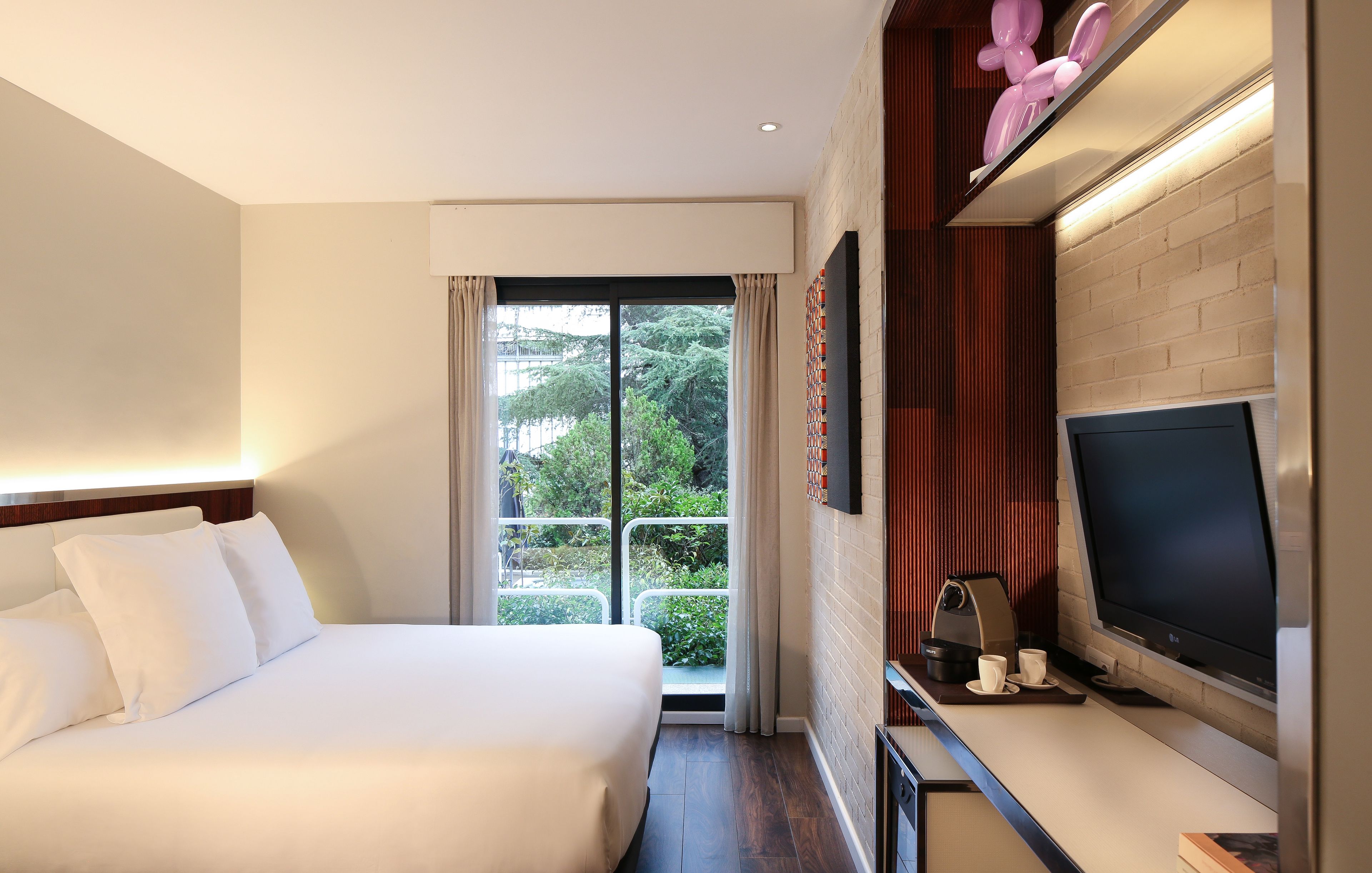Habitación de hotel con cama, televisión, cafetera y balcón con vistas a zonas verdes.