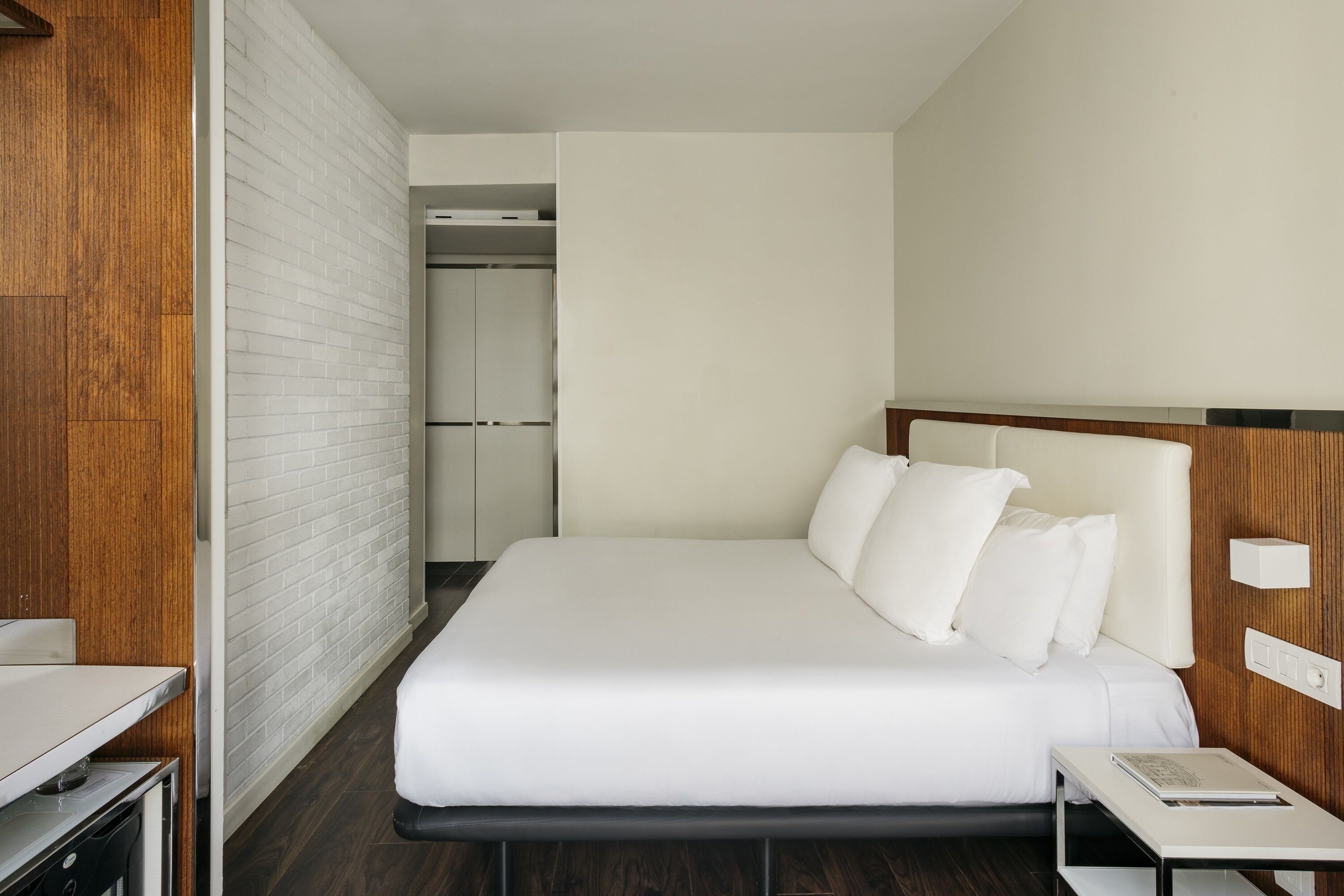 Habitación de hotel con una cama grande, cabecera de madera, mesita de noche y pared blanca.