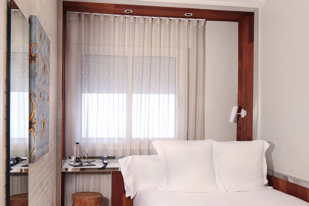 Une chambre d’hôtel comprenant un lit, un bureau avec une chaise, un tabouret en bois, un miroir et une fenêtre avec des rideaux.