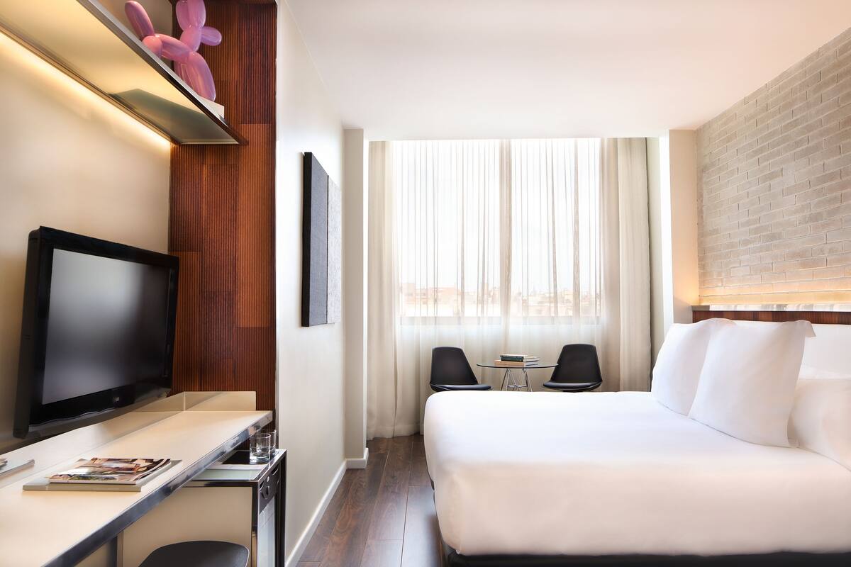 Une chambre d’hôtel comprenant un lit, un bureau avec un téléviseur à écran plat, une petite table, deux chaises et une fenêtre avec des rideaux légers.