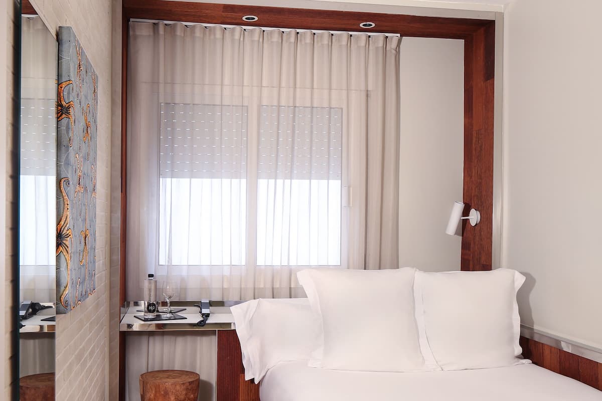 Une chambre d’hôtel comprenant un lit, un bureau, une chaise, un tabouret en bois, un miroir et une fenêtre avec des rideaux.