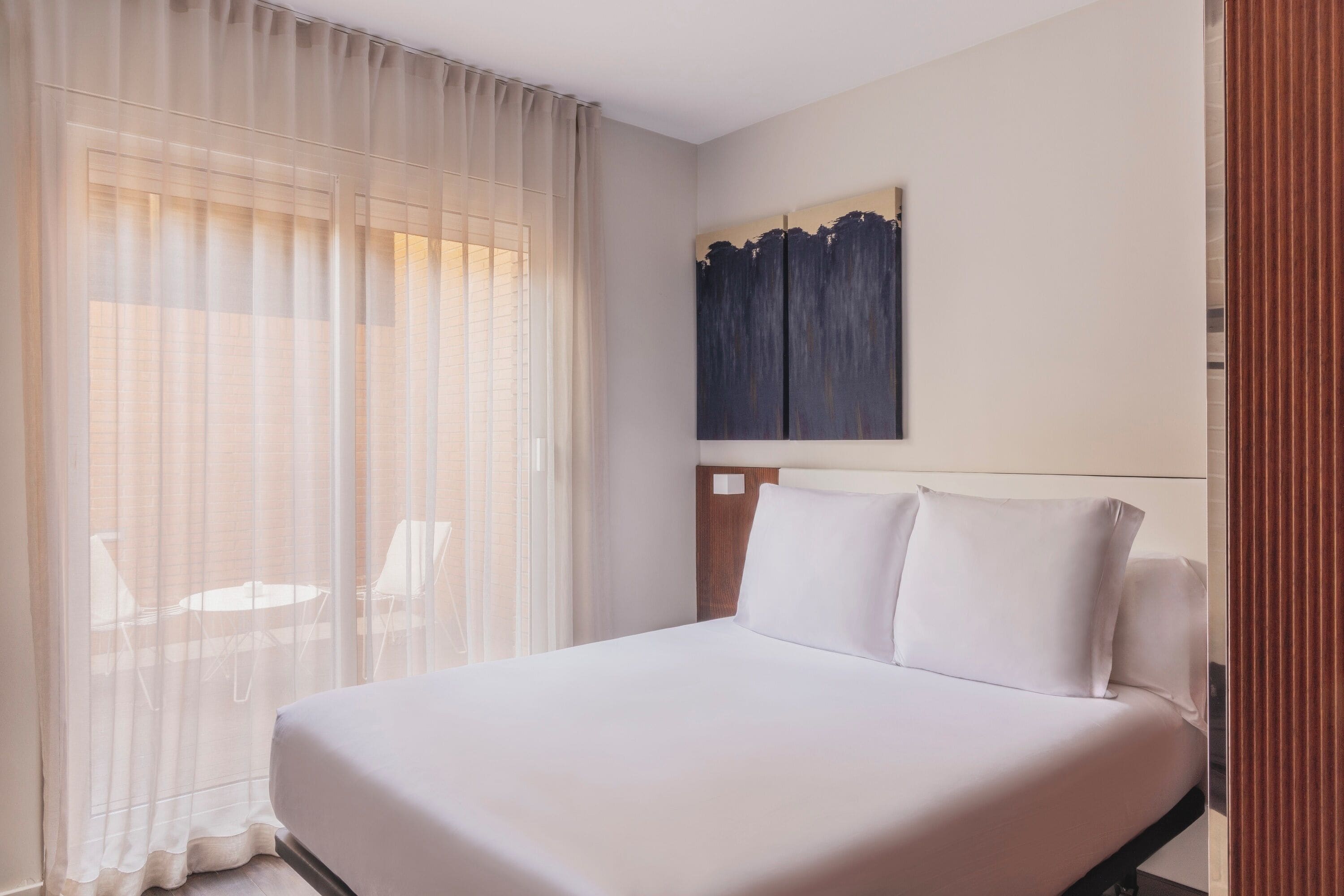 Habitación de hotel con cama, una ventana grande con cortinas translúcidas, un cuadro en la pared y cabecera de madera.