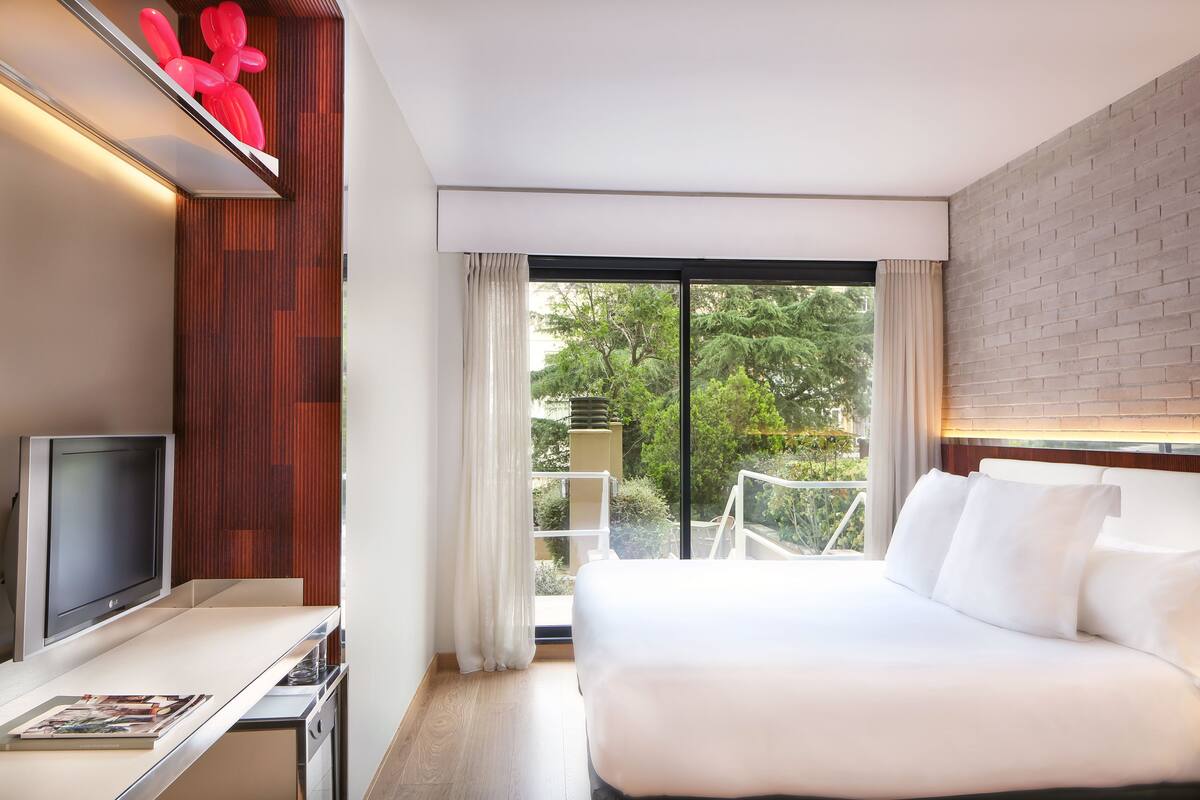 Une chambre d’hôtel moderne dotée d’un grand lit, d’une télévision à écran plat, d’un petit bureau et d’un balcon offrant une vue sur les arbres.