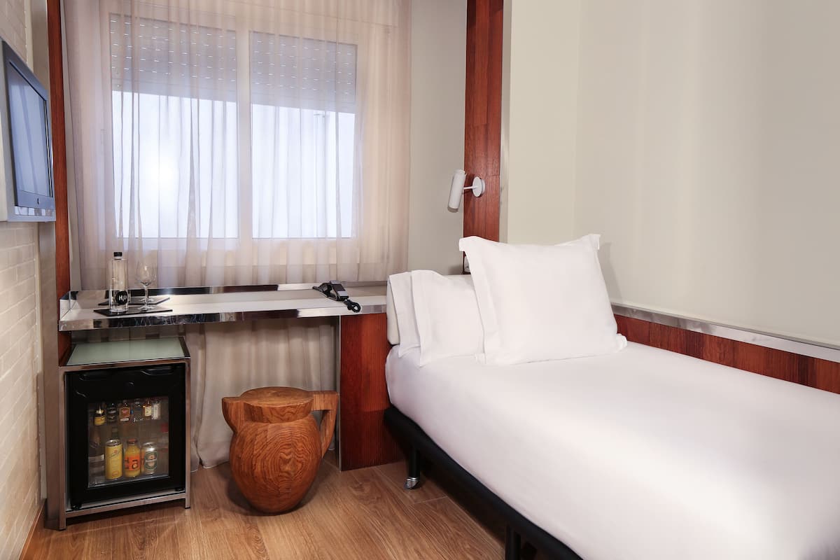 Une chambre d’hôtel compacte comprenant un lit, un petit bureau avec un ordinateur, un minibar et un tabouret en bois.