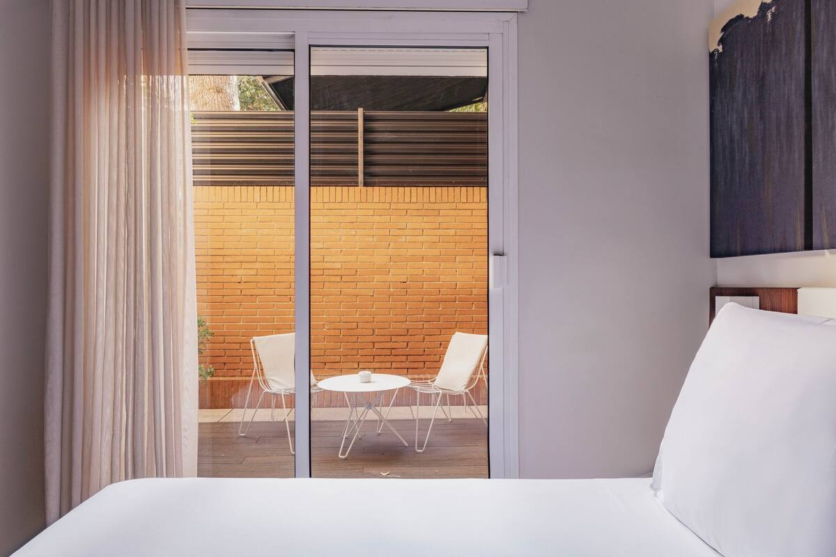 Une chambre d’hôtel avec un lit, une porte-fenêtre coulissante donnant sur une terrasse extérieure avec un mur en briques, une table ronde et deux chaises.