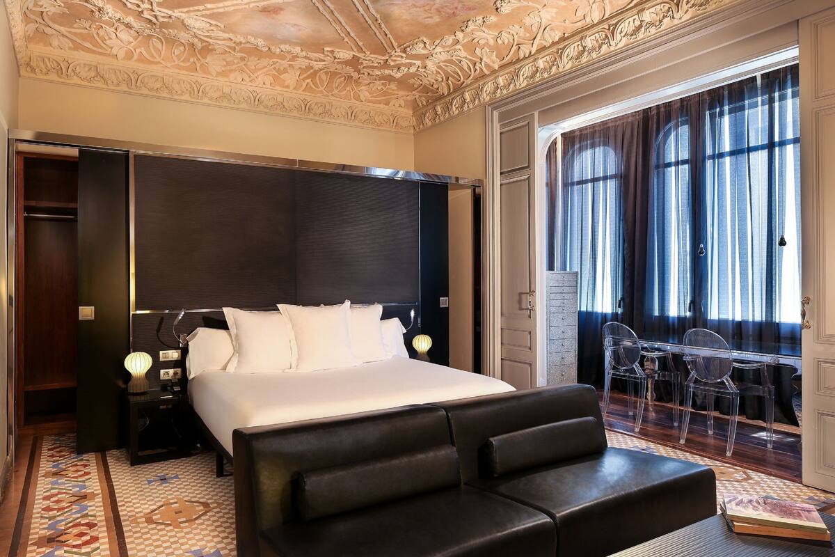 Une chambre d’hôtel avec un grand lit, une table de chevet, un bureau avec une chaise et un balcon avec des rideaux.