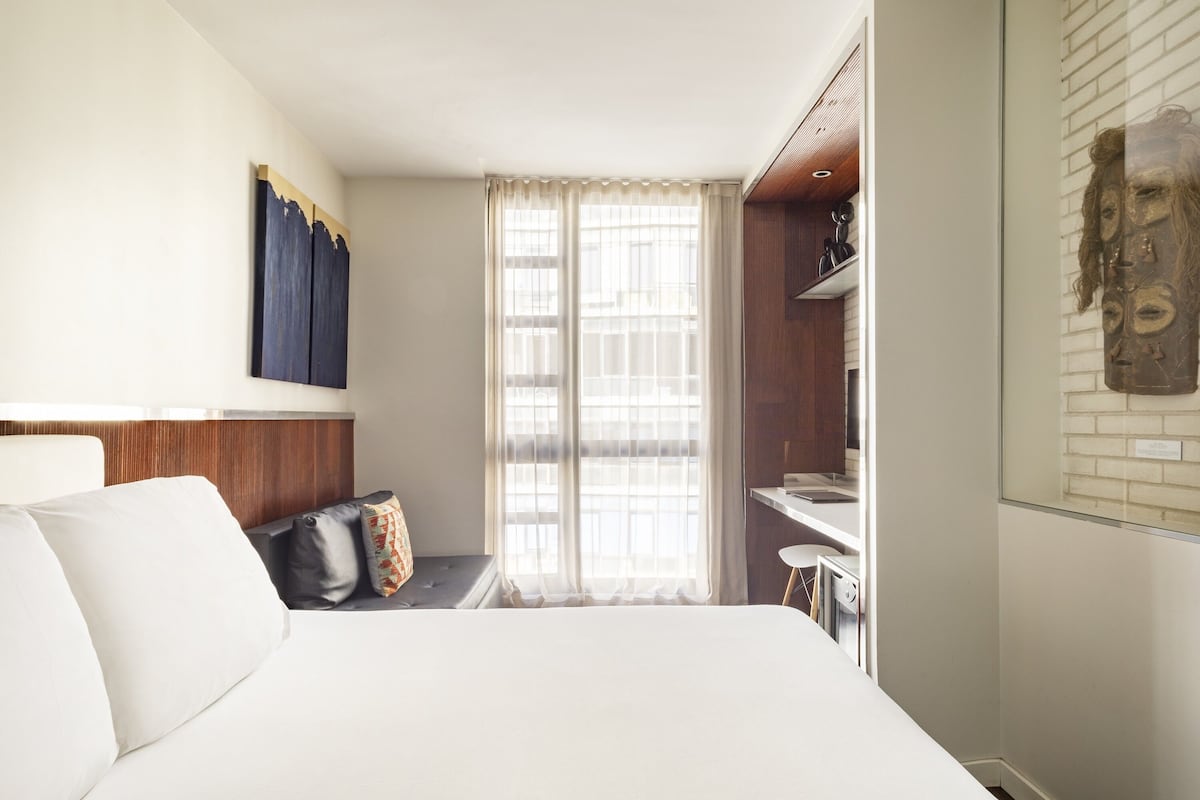 Une chambre d’hôtel avec un lit, une fenêtre avec des rideaux, un petit bureau et une œuvre d’art accrochée au mur.