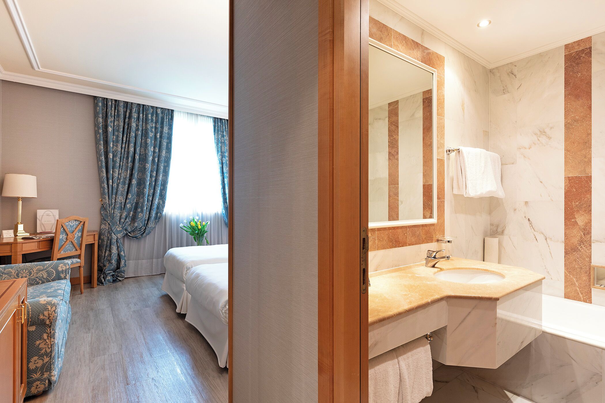 A hotel room with a bed, a desk, a chair, a large mirror, and a bathroom with a sink and bathtub.